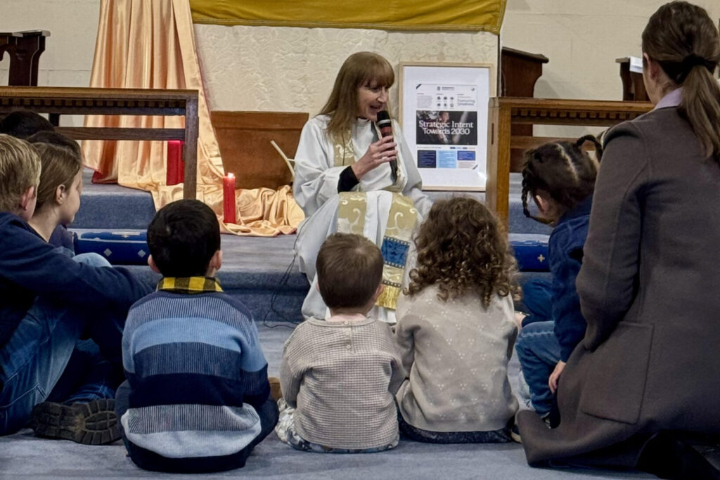A group of children sit in a circle at the front of the Church and Rev'd Rachel (centre) gives Children's Talk